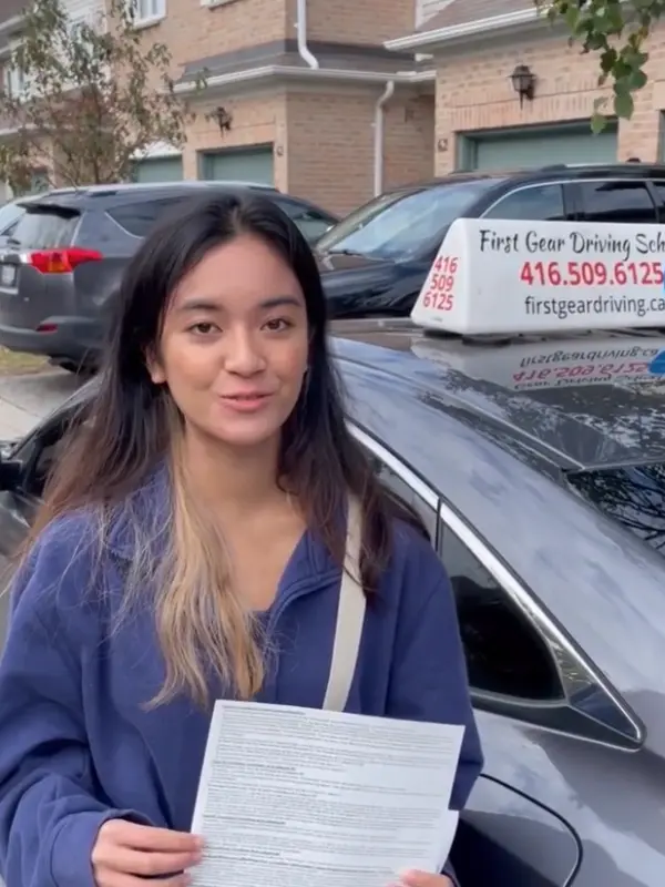 A First Gear student holding her road test result beside the First Gear car with its roof sign